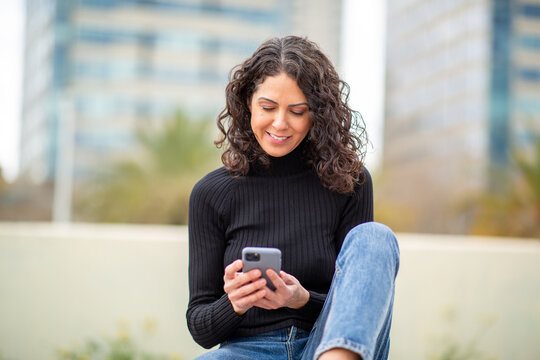 young woman with curly hair sits outdoors in an urban setting, smiling as she looks at her smartphone