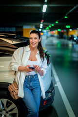 Brunette haired woman standing next to her car in parking garage and using smart phone