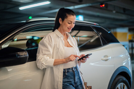 Brunette haired woman standing next to her car in parking garage and using smart phone