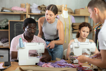 Friendly skilled young seamstress mentoring fashion students in clothing construction class,...