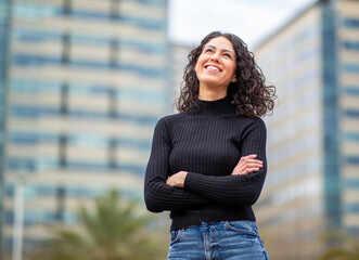 woman stands outdoors with arms crossed smiling and looking upward with city buildings in the...