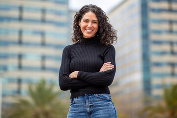 confident woman with curly hair smiles while standing with her arms crossed in front of city buildings.