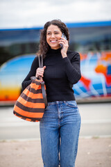 Woman carrying bag and smiling while talking on the phone carrying bag, in front of bus