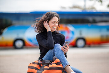 Travel woman sitting outdoors smiling at her phone with bus in the background.