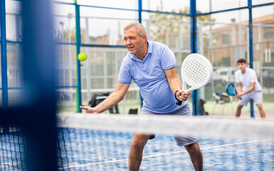 Focused aged man playing friendly paddleball match on outdoor summer court. Senior people sports concept..