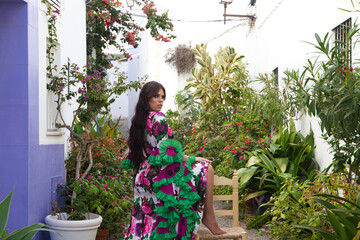 Young and beautiful woman dancing flamenco rests one foot on an old wooden chair in a typical street of andalusia with flowers. She is dressed in a tailed gown with ruffles and flowers.