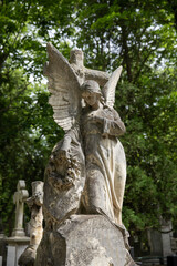 Weathered stone angel statue with wings and a wreath in an old cemetery. Surrounded by green trees and gravestones. Peaceful, somber atmosphere with historic and spiritual symbolism.