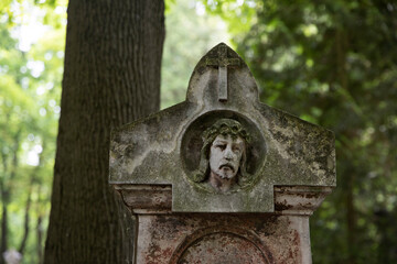 Old weathered gravestone with a carved face of Jesus Christ crowned with thorns and a cross above. Surrounded by forest trees. Symbol of faith, sorrow, and remembrance in a cemetery.