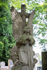 Weathered stone angel statue with wings and a wreath in an old cemetery. Surrounded by green trees and gravestones. Peaceful, somber atmosphere with historic and spiritual symbolism.