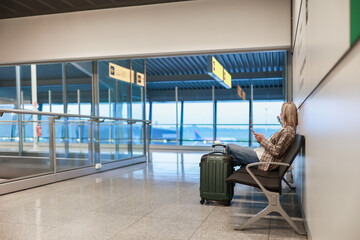 A young woman sits sideways in an airport terminal, with a suitcase beside her, quietly waiting for...