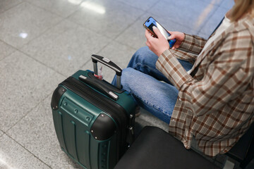 A young woman sits in the airport holding her phone and suitcase, checking her travel details in a...