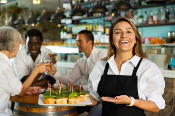 Cheerful smiling female waiter holding served tray meeting visitors at comfortable bar