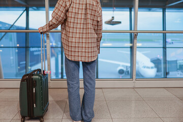 A traveler stands with their back to the camera, holding a suitcase near the airport window...
