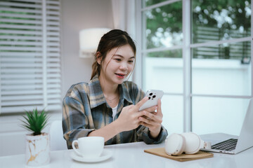 A young woman wearing headphones smiles while using her smartphone at home. Surrounded by cozy...