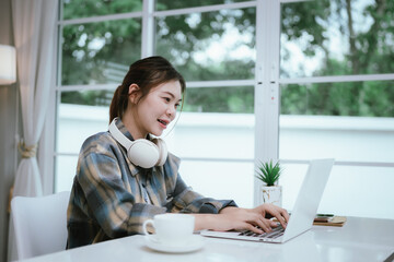 A young woman with long hair wears headphones while working or studying on a laptop at home,...