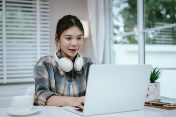 A young woman with long hair wears headphones while working or studying on a laptop at home,...