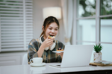 A young woman enjoys coffee and dessert while wearing headphones, working or studying remotely from...
