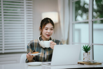 A young woman enjoys coffee and dessert while wearing headphones, working or studying remotely from...