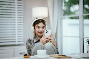 A young woman wearing headphones smiles while using her smartphone at home. Surrounded by cozy...