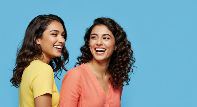 Two young beautiful women laughing happily together on blue background