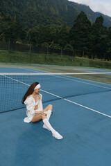 Young female tennis player in sporty outfit sitting on the court, expressing relaxation and...