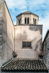 Ancient stone bell tower rises above terracotta rooftops in Girona, Spain. A narrow alley with beige walls, tiled roofs, and a grilled window highlights traditional Mediterranean architecture