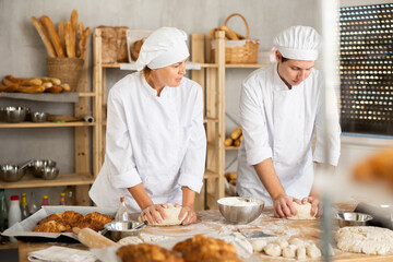 Two skilled female and male bakers in white uniform kneading dough and preparing portioned pieces before baking in small bakehouse