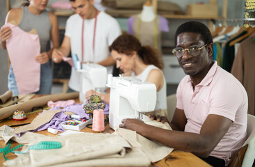 Positive middle-aged man sewing while others working with sewing-machine and making details in...