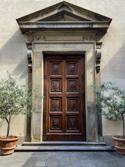 ornate wooden door with carved panels in a stone archway, flanked by potted olive trees against a beige wall. old