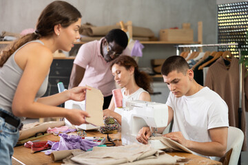 During lesson, African male teacher helps student sew, while others work with fabric, blanks, and patterns. Interest group, activities, hobbies, vocational training