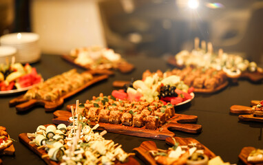 Luxurious catering table with assorted appetizers, fruit platters, and gourmet bites on wooden boards, beautifully lit for an elegant event or celebration.