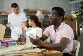 Middle-aged man of African descent stitching at machine and speaking with female classmate at table during beginner sewing workshop while other students handling fabric, paper patterns, and mannequin