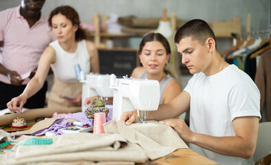 Focused young guy working at sewing machine during hands-on course and talking to classmates preparing patterns, fabric and fitting garments..