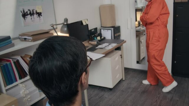 Handheld shot of young female stressed prisoner in orange jumpsuit talking to mature psychotherapist analyzing criminal profile during therapy session