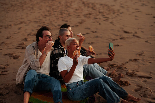 Friends eating donuts and taking a photo of themselves on beach at sunset - Powered by Adobe