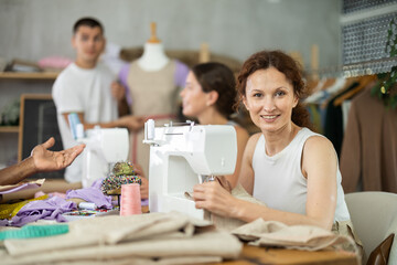 Attentive middle-aged woman sewing while others working with sewing-machine and mannequin in dressmaking courses