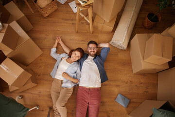 A couple lies on the floor of their new home, smiling and relaxed, surrounded by packed moving...