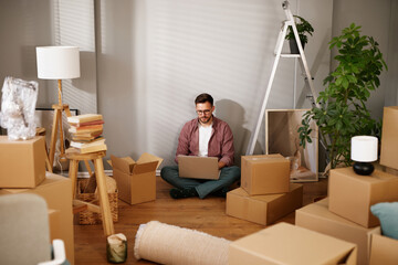 A person sits on the floor amidst unpacked cardboard boxes, using a laptop to organize tasks after...