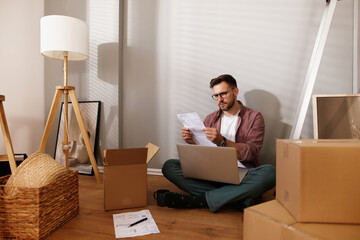 A person sits on the floor of a new home surrounded by cardboard boxes and a laptop, reviewing...