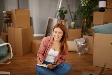 Woman sits on the floor of her new home, writing notes and organizing her thoughts amid cardboard...