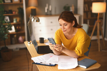 A woman wearing a yellow top looks at her smartphone while leaning over a cluttered desk in a...