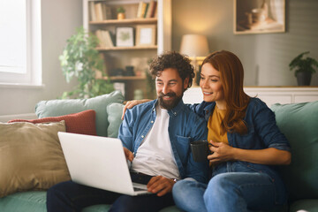 A couple sits closely on a sofa, engaged with a laptop. They share smiles and laughter, surrounded by a warm living room ambiance filled with books and plants.
