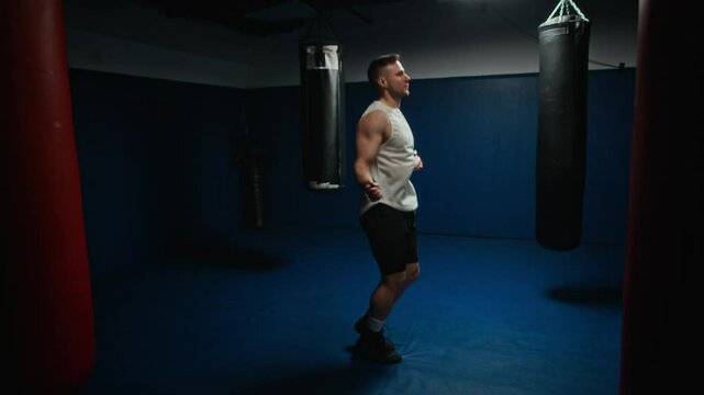 Full shot of athletic male boxer sharpening his footwork and conditioning by jump roping between heavy bags in dark, focused gym environment, preparing for peak performance