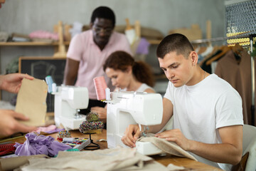 Engaged young man enjoying hands-on learning during group sewing class for adults, stitching at machine in tailoring studio, while fellow learners sharing creative work and friendly conversation..