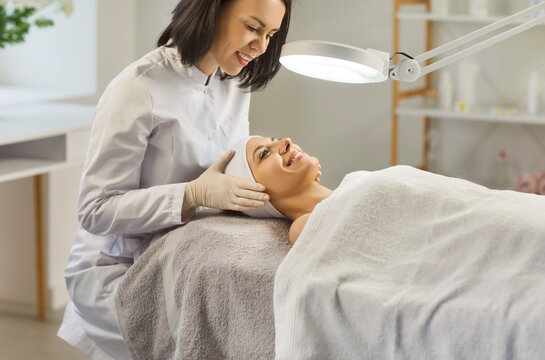Cosmetologist examining face of young smiling woman lying on couch under bright lamp. Friendly skincare specialist evaluating skin condition before starting professional beauty procedure in spa. - Powered by Adobe