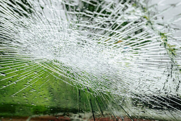 A cracked car windshield seen from the inside, with raindrops and a blurred outdoor scene including...