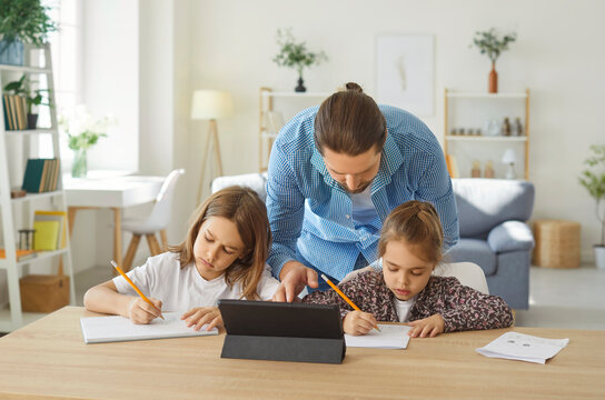 Loving caring father helps children with school homework. Two creative kids sitting at desk with tablet device, having remote class, training writing skills and drawing pictures while dad is watching