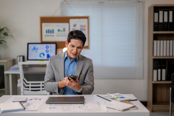 A man in a suit is looking at his cell phone while sitting at a desk with papers