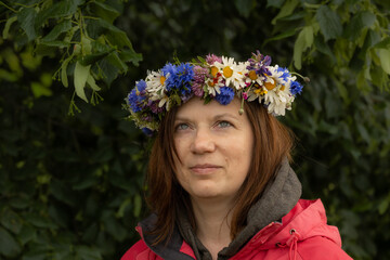 Woman with light eyes and reddish-brown hair wears a colorful wildflower crown featuring daisies,...
