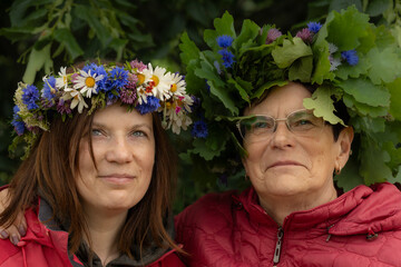 Two women wearing handmade floral wreaths smile warmly, dressed in red jackets and standing close...